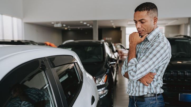 A man stands inside a car dealership  and contemplates the price of a vehicle. 